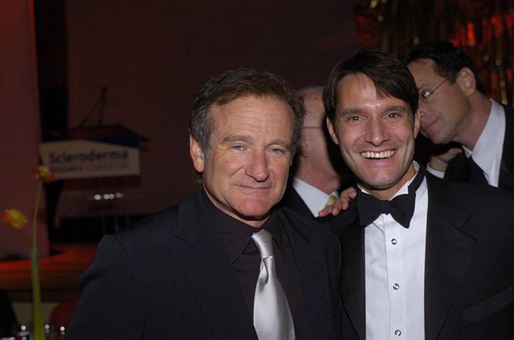 Two men in formal attire are smiling at an event. The man on the left is wearing a suit and tie, while the man on the right is dressed in a tuxedo. A podium is visible in the background.