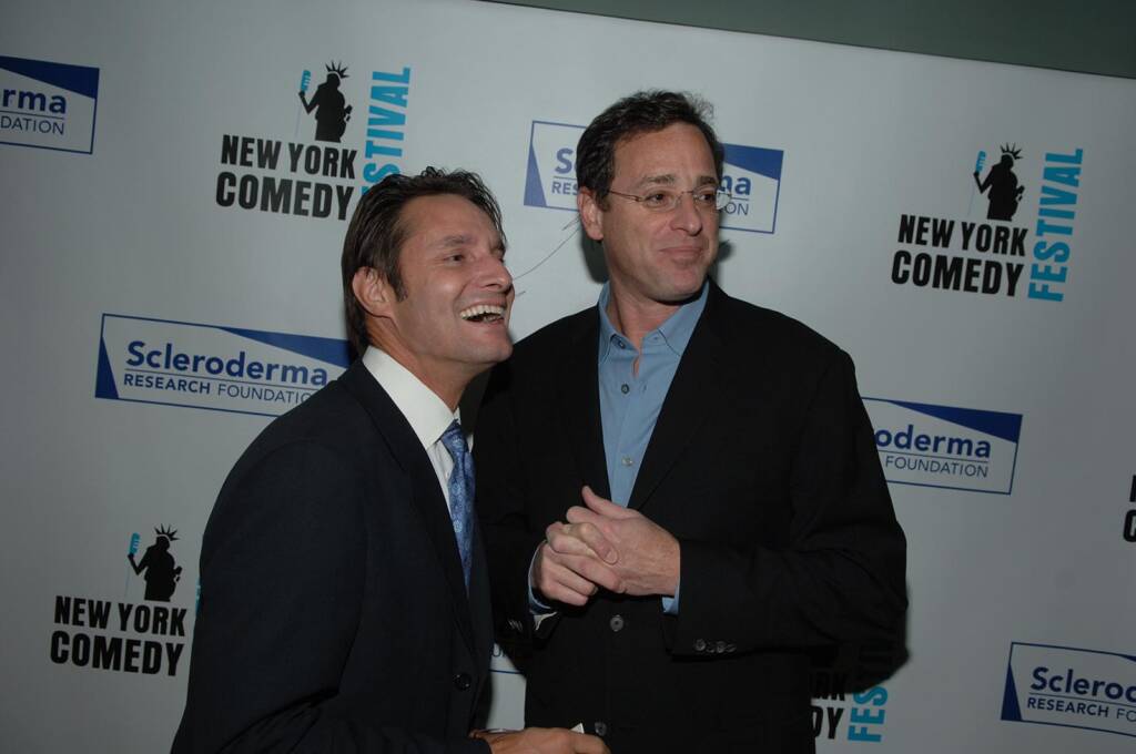 Two men in suits stand in front of a backdrop with logos for the Scleroderma Research Foundation and the New York Comedy Festival. One man is laughing while the other is looking to the side.