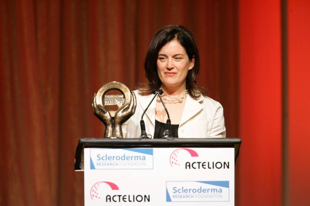 A woman in a white blazer stands at a podium holding an award, with banners for the Scleroderma Research Foundation and Actelion behind her.