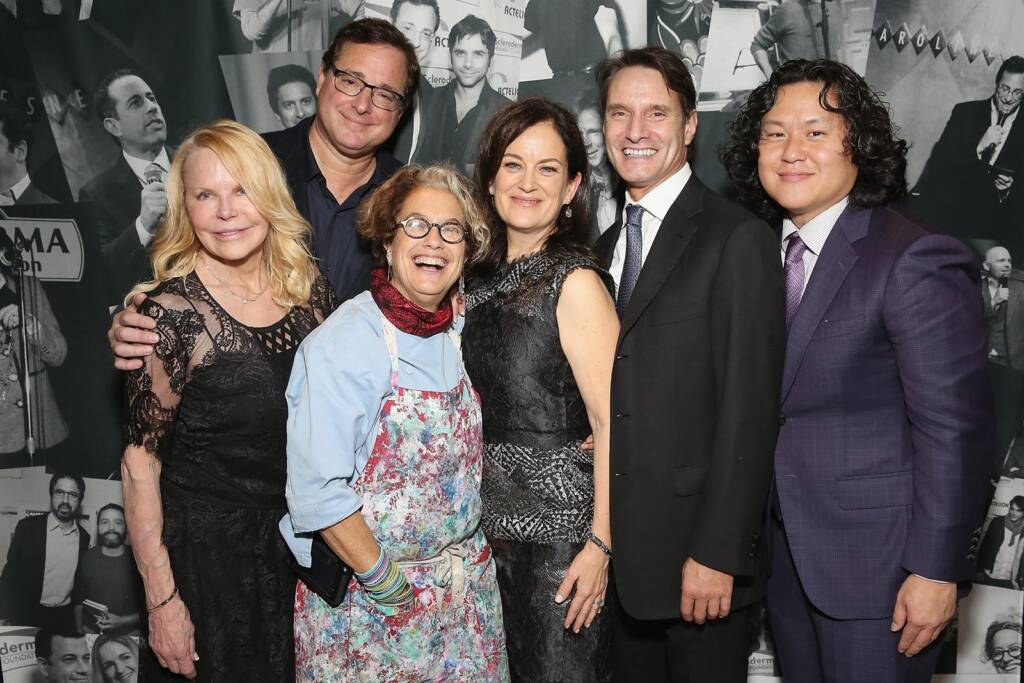 Six individuals are standing close together, dressed in various formal and casual outfits, smiling at the camera against a backdrop with multiple photographs.