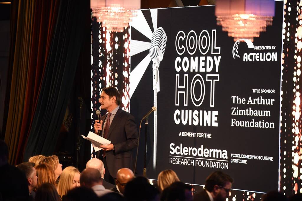 Luke Evnin speaks into a microphone on stage at the "Cool Comedy Hot Cuisine" event, a benefit for the Scleroderma Research Foundation, sponsored by Actelion and The Arthur Zimtbaum Foundation.